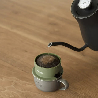 Coffee being poured from a black kettle into a green coffee filter resting on a titanium mug on a wooden surface.