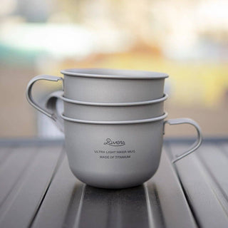 Two silver mugs stacked on a reflective surface with a blurred outdoor background